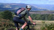 Man riding a gravel bike wearing a green jersey, black shorts and a green gilet with a pink and blue rucksack on